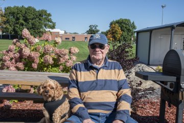 David Dubbelde in cap, orange and blue striped shirt, and sunglasses sits with his dog sit on a bench at Homecoming 2026 Construction Management BBQ.