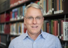Herb LaGoy in glasses and blue button down shirt stands in front of library stacks.