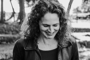 Black and White image of Jessica Cuello in leather jacket, smiling, walking through woodded area.