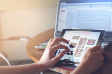 Hands hold an iPad while a laptop screen behind it shows email open.