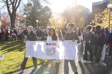 President Nesbitt, student Madison Jordan, and Director of Community, Belonging and Engagement Kira Maddox carry a banner as a crowd of students, faculty, and staff follow behind them.