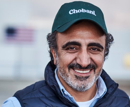 Hamdi Ulukaya, founder and CEO of Chobani, in a Chobani hat and blue vest, smiles at the camera.