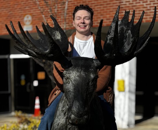 Marshall Hildreth '26 sits on top of a moose sculpture outside a brick building.