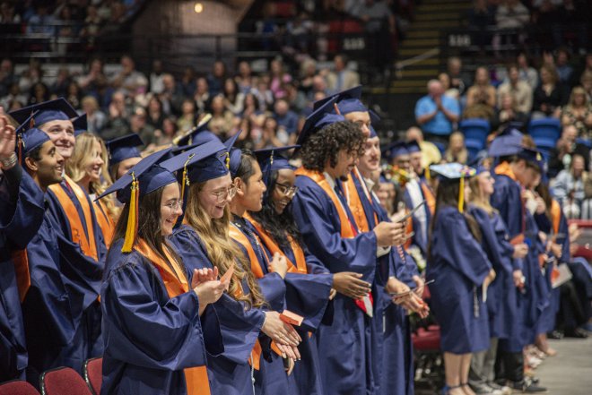 A row of graduates in blue commencement gowns and caps in an auditorium.