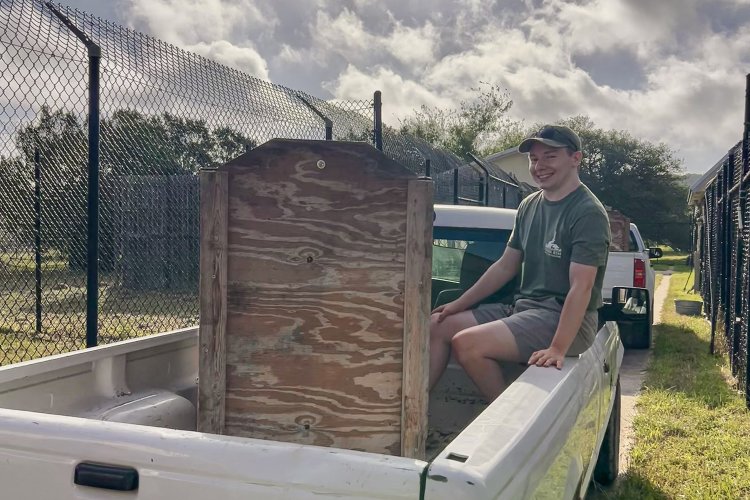 Marshall Hildreth in shorts, green t-shirt, and hat, sits in the back of a pick-up truck.