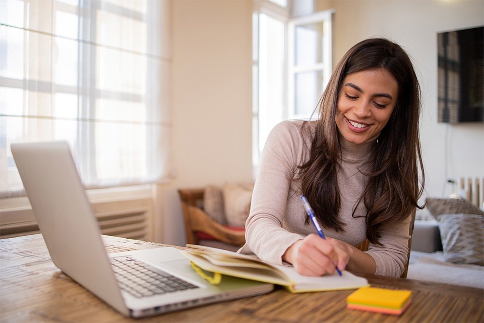 Female representing person with long, brown hair at laptop computer taking notes in a yellow notebook. She is sitting in a home living room presumably taking notes to compare different online business degrees.